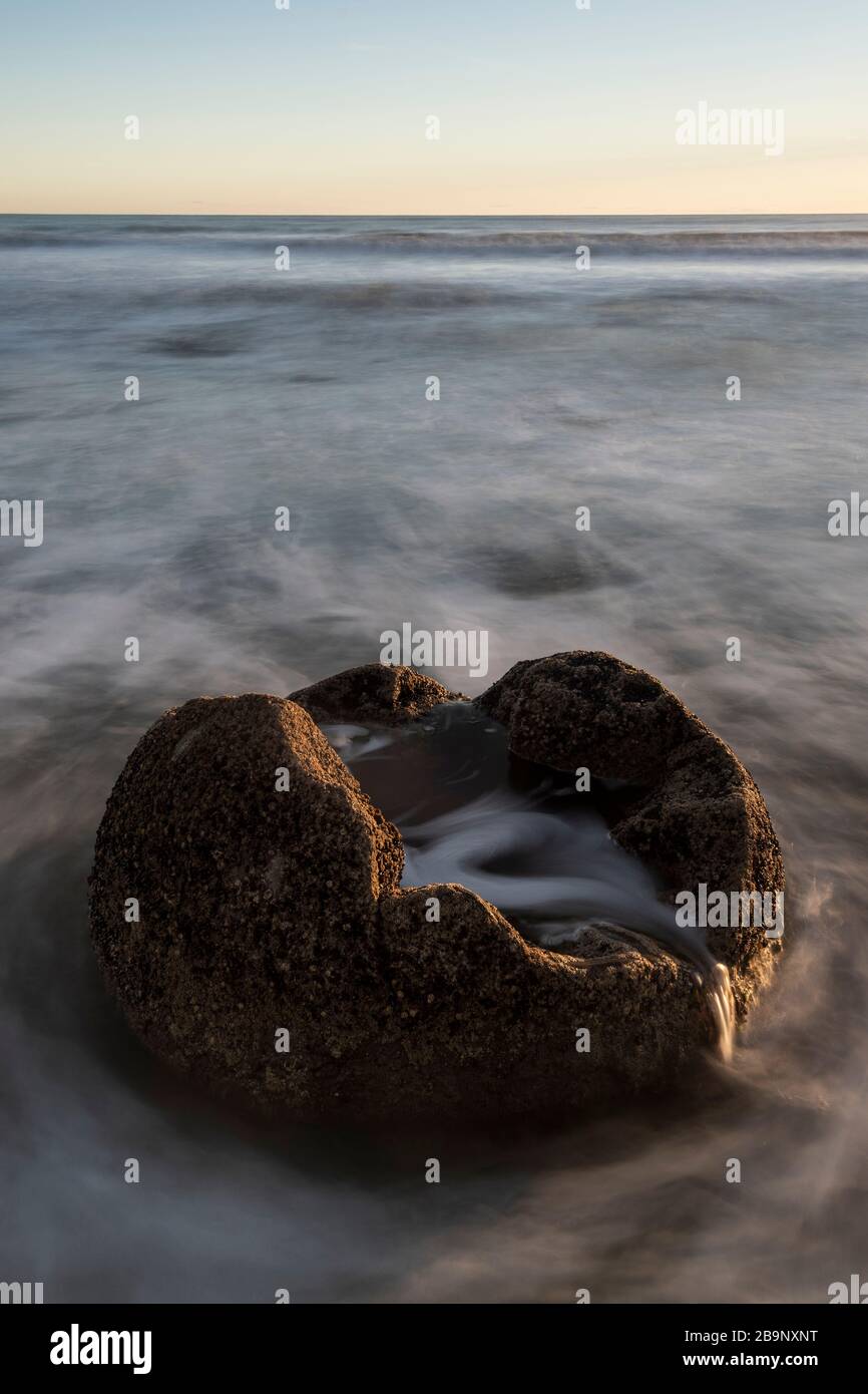 Einer der Moeraki Boulders bei Sonnenaufgang mit einem farbenfrohen Himmel, Neuseeland. Der Felsbrocken scheint von dem in Bewegung befindlichen Meerwasser und dem Himmel in ora umgeben Stockfoto