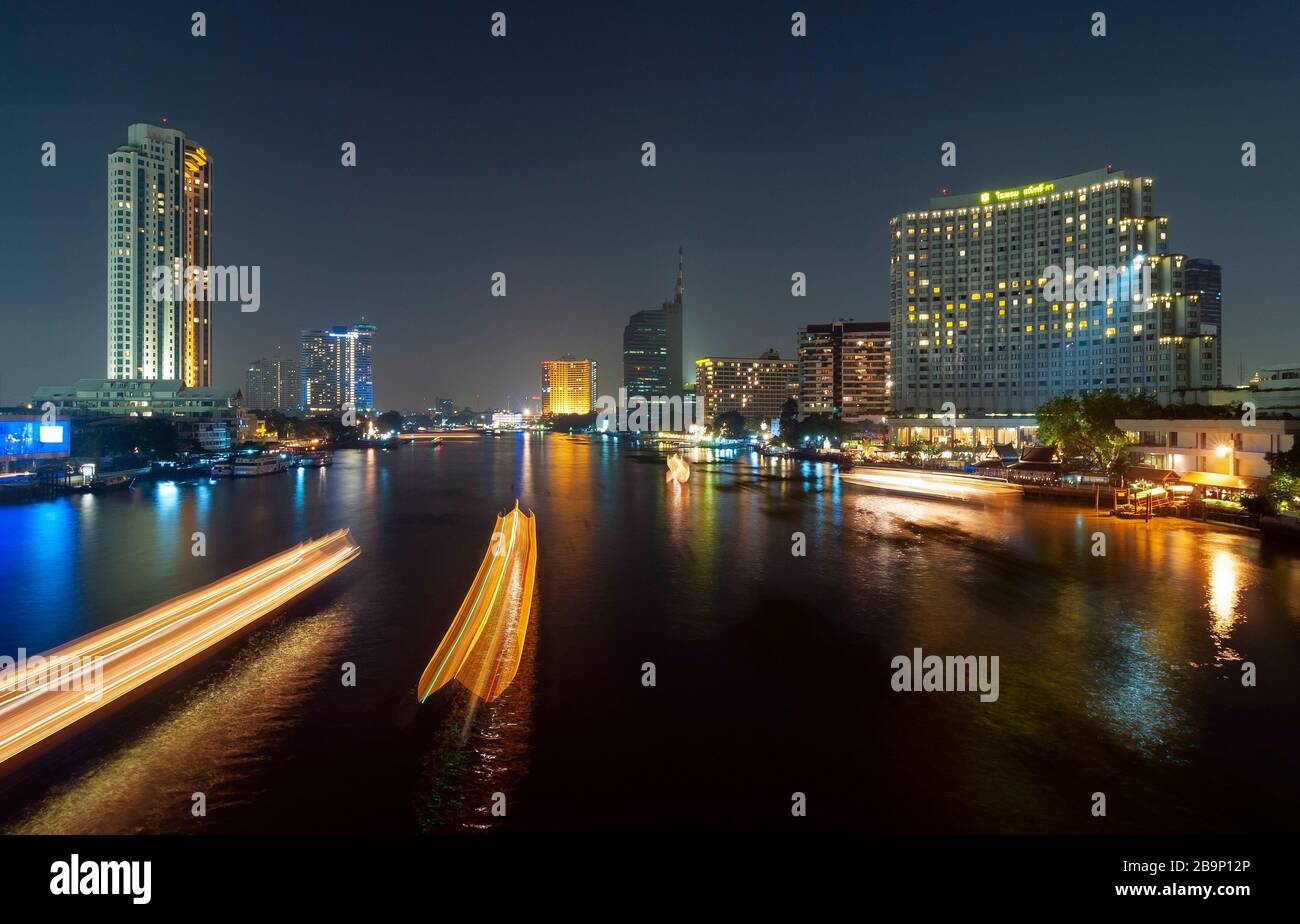Langzeitfotografie des Chao-Phraya-Flusses mit verschwommenen Bewegungen beleuchteter Boote und der städtischen Skyline von Bangkok in der Nacht, Thailand. Stockfoto