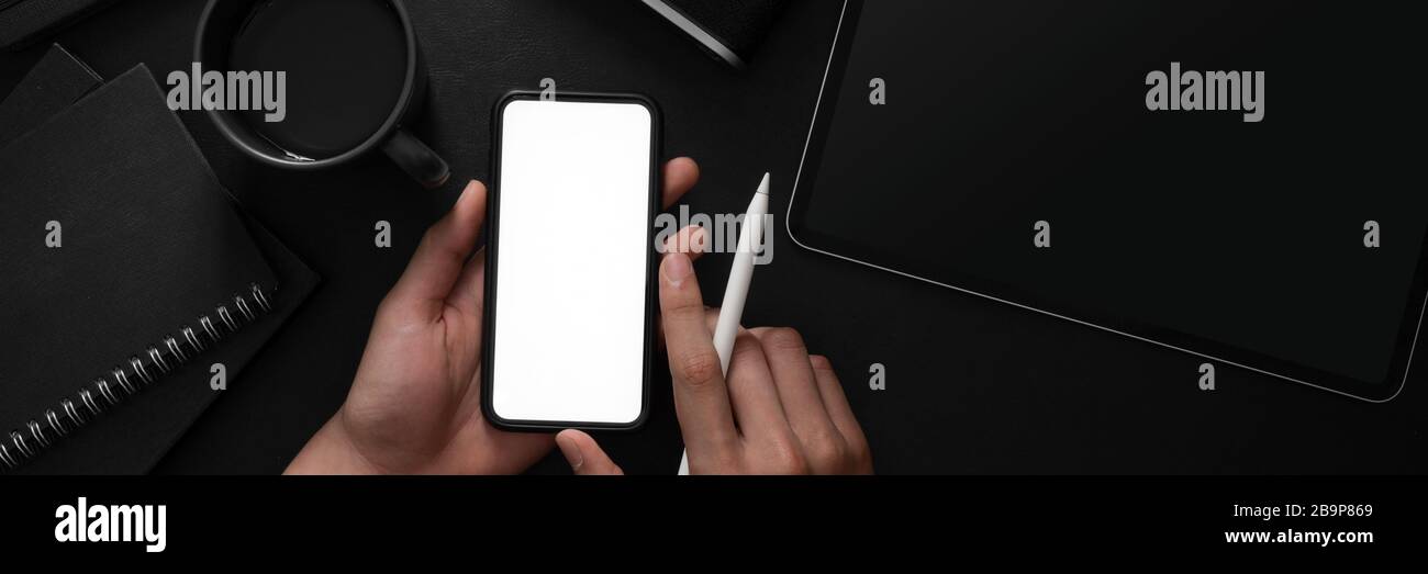 Overhead-Aufnahme eines Mannes, der ein leeres Bildschirm-Smartphone in einem dunklen, modernen Arbeitsbereich mit anderen Büromaterialien hält Stockfoto
