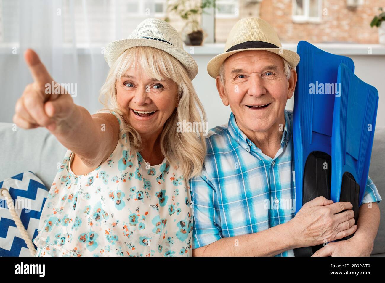 Senioren Paar sitzt im Reisebüro sitzen mit Hüten Mann hält Flossen Frau zeigt Kamera aufgeregt Stockfoto