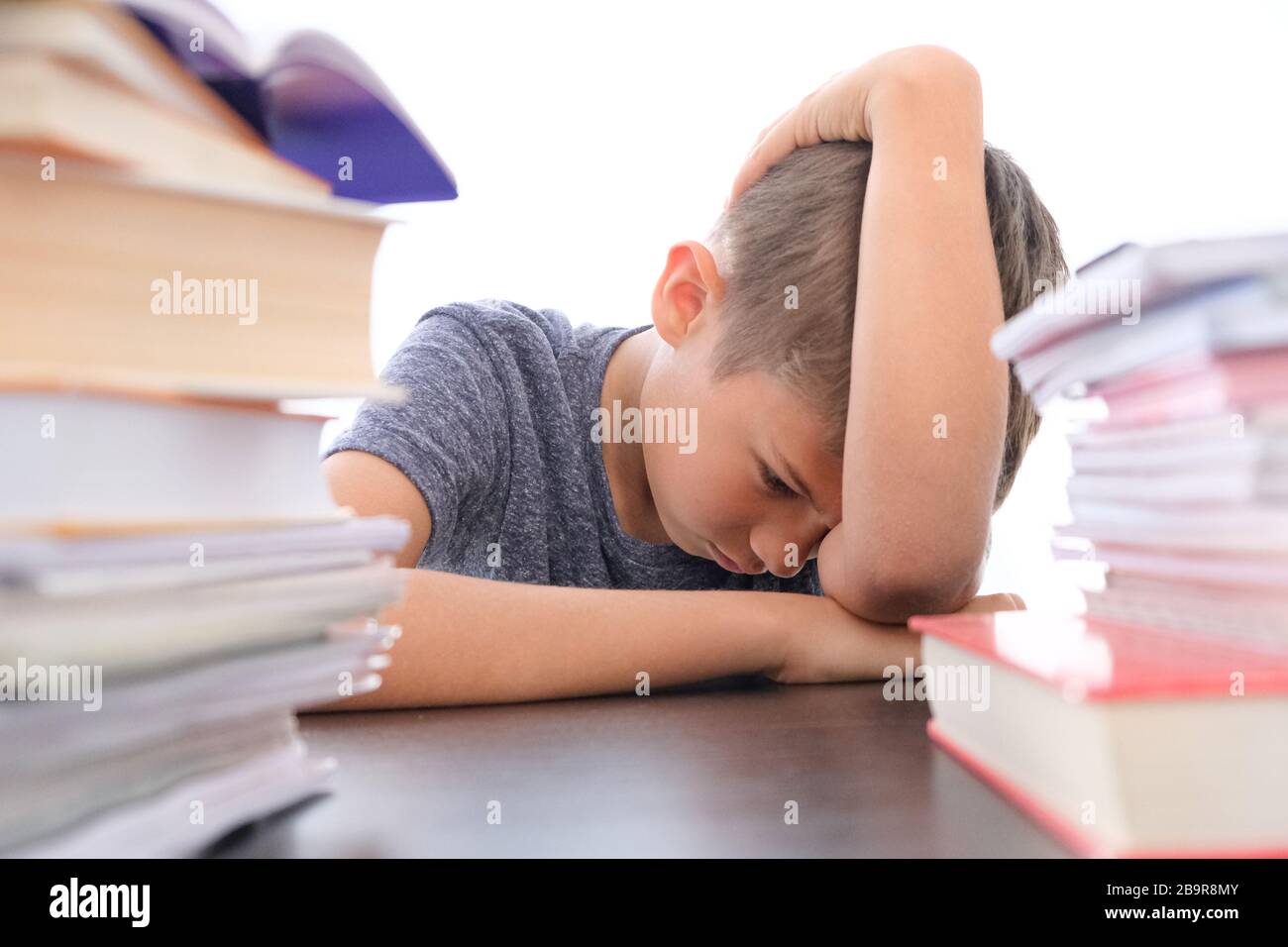 Müde enttäuschte der Schuljunge, der seinen Kopf zwischen den Haufen von Büchern, Lehrbüchern und Schulbüchern auf seinem Schreibtisch zu Hause sitzend senkte Stockfoto