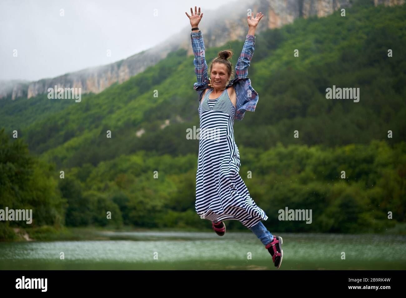 Locker gekleidete Wanderfrau unter dem Regen springt in der Nähe des Sees in Bergen. Ehrwürdige und authentische Ausdrucksweise. Stockfoto