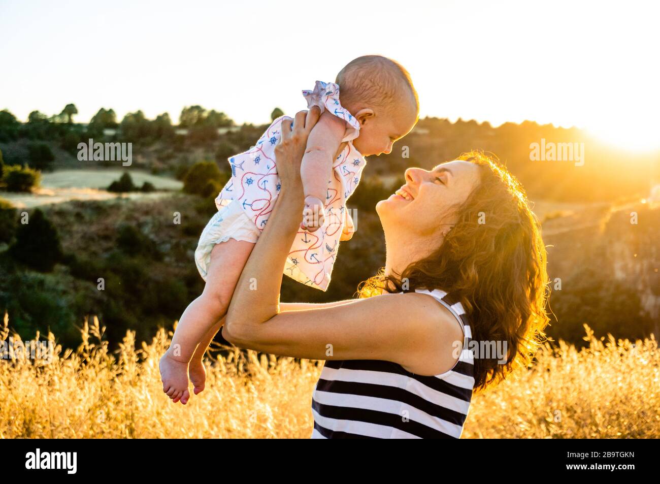 Glückliche Mutter hebt Baby zur goldenen Stunde mitten auf den Feldern in die Luft Stockfoto