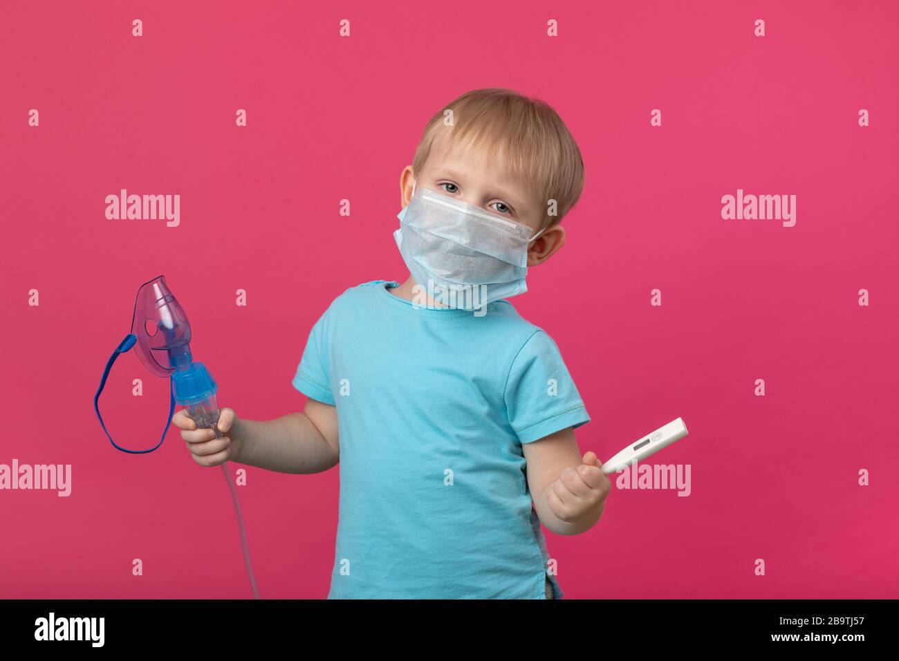 Ein blondes Kind mit Inhalator und Thermometer in der Hand auf einem einfarbig rosafarbenen Hintergrund. Studio-Fotografie zur Behandlung von Temperatur, Husten und Stockfoto