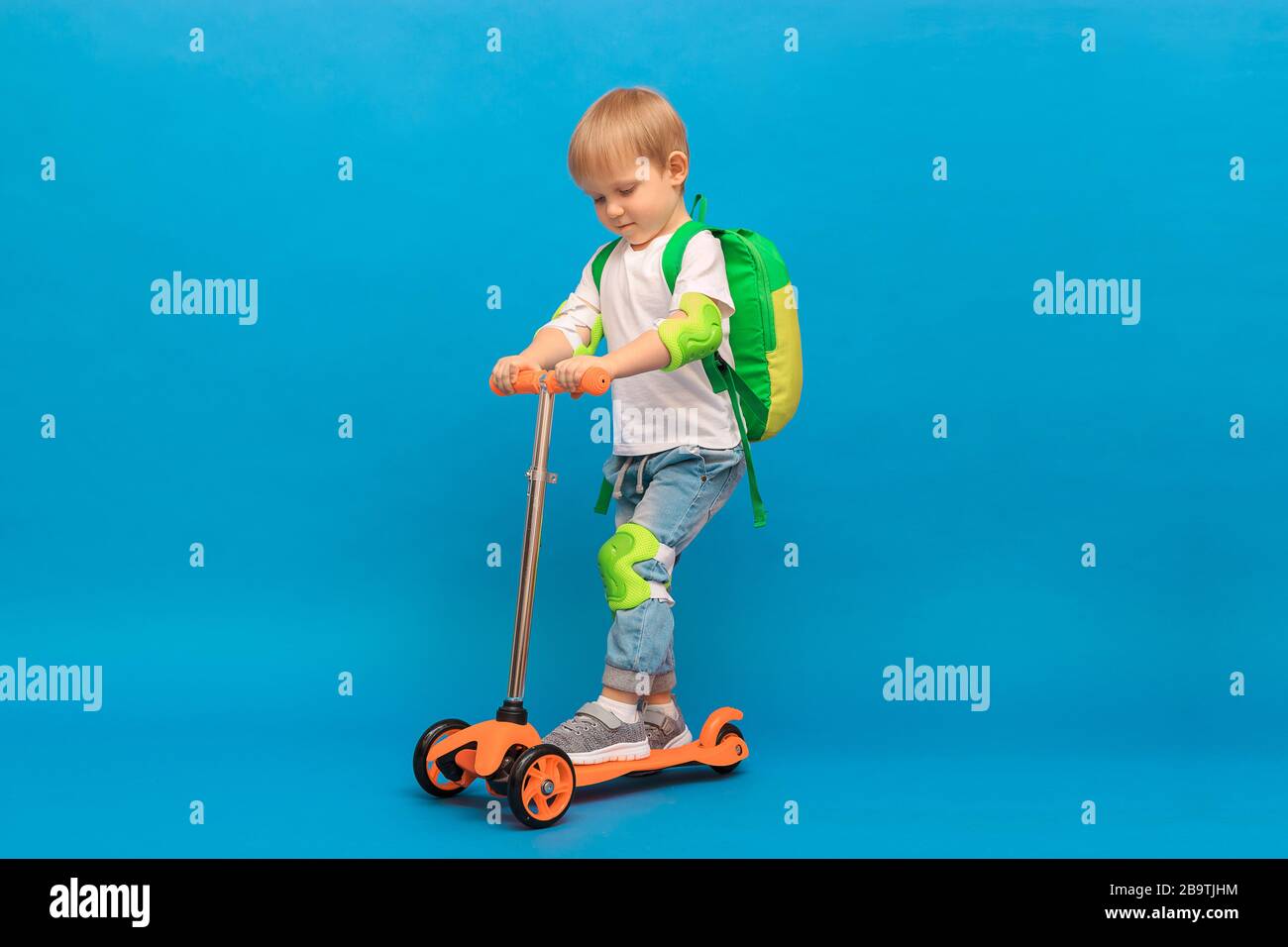 Ein 4-jähriger blondhaariger Junge, ein Kind mit Rucksack und in Jeans mit weißem T-Shirt reitet auf einem orangefarbenen Roller mit hellgrünem Schutz auf Stockfoto