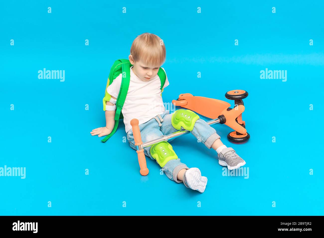 Der blonde Junge, der mit einem Rucksack auf Knien und Ellbogen geschützt ist, ist von einem Roller auf hellblauem Hintergrund gefallen. Studio Shot Konzept für Advertisi Stockfoto