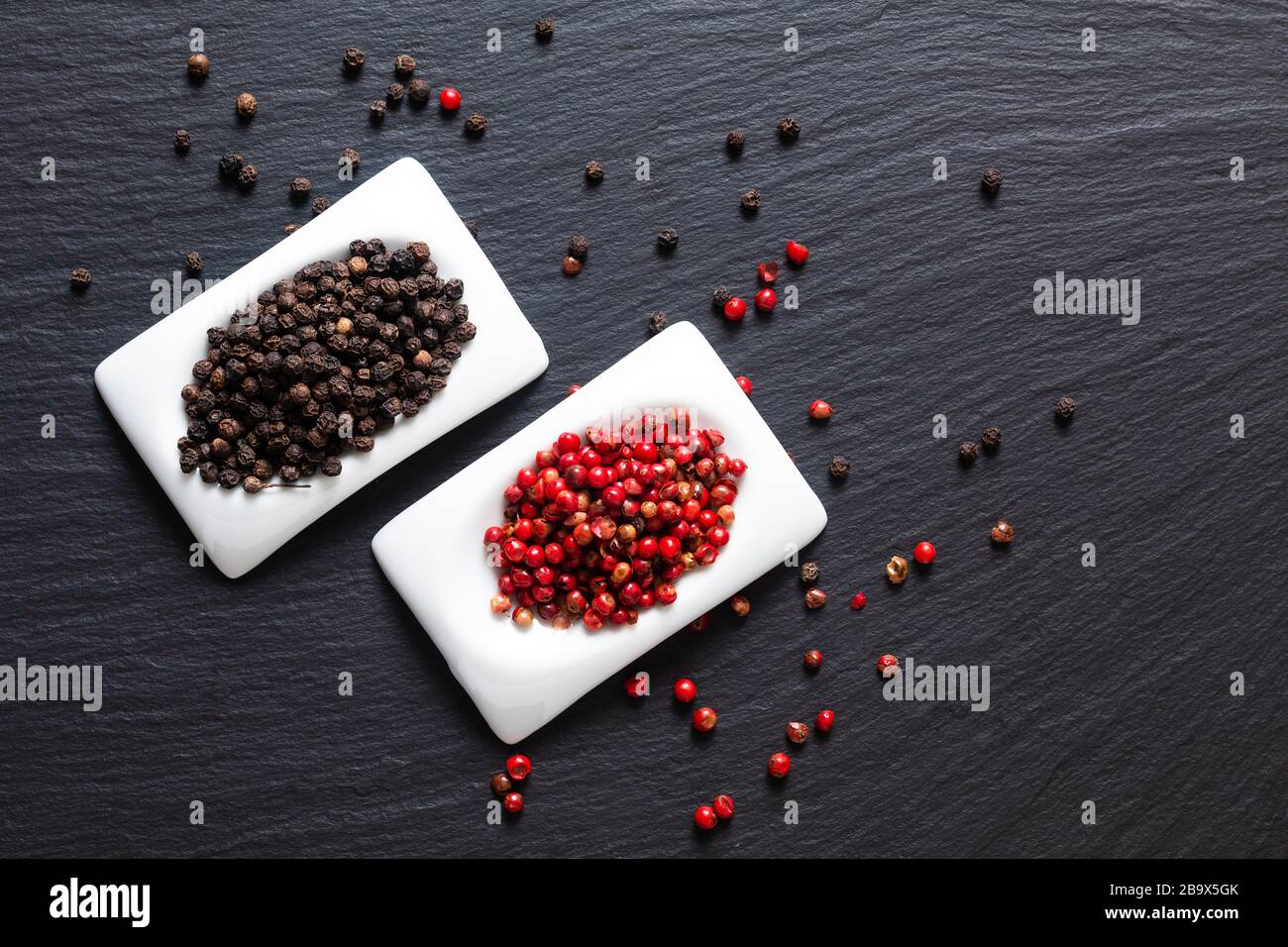 Konzept für gesunde Lebensmittelzutaten organische schwarze Paprika und rote Pfefferkörner in quadratischem Keramikbecher auf schwarzem Schiefersteinbrett mit Kopierfläche Stockfoto