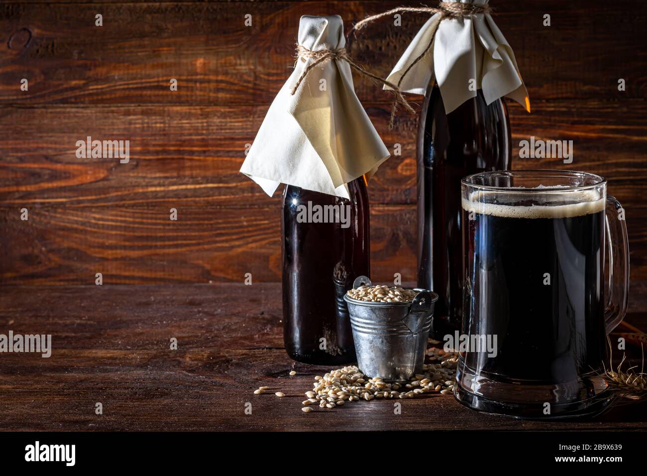 Hausgemachtes dunkles Bier in Flaschen und ein Glas auf dunklem Hintergrund. In der Nähe liegen Gerstenkörner und Weizenkörner. Dunkler Hintergrund. Kopierbereich Stockfoto