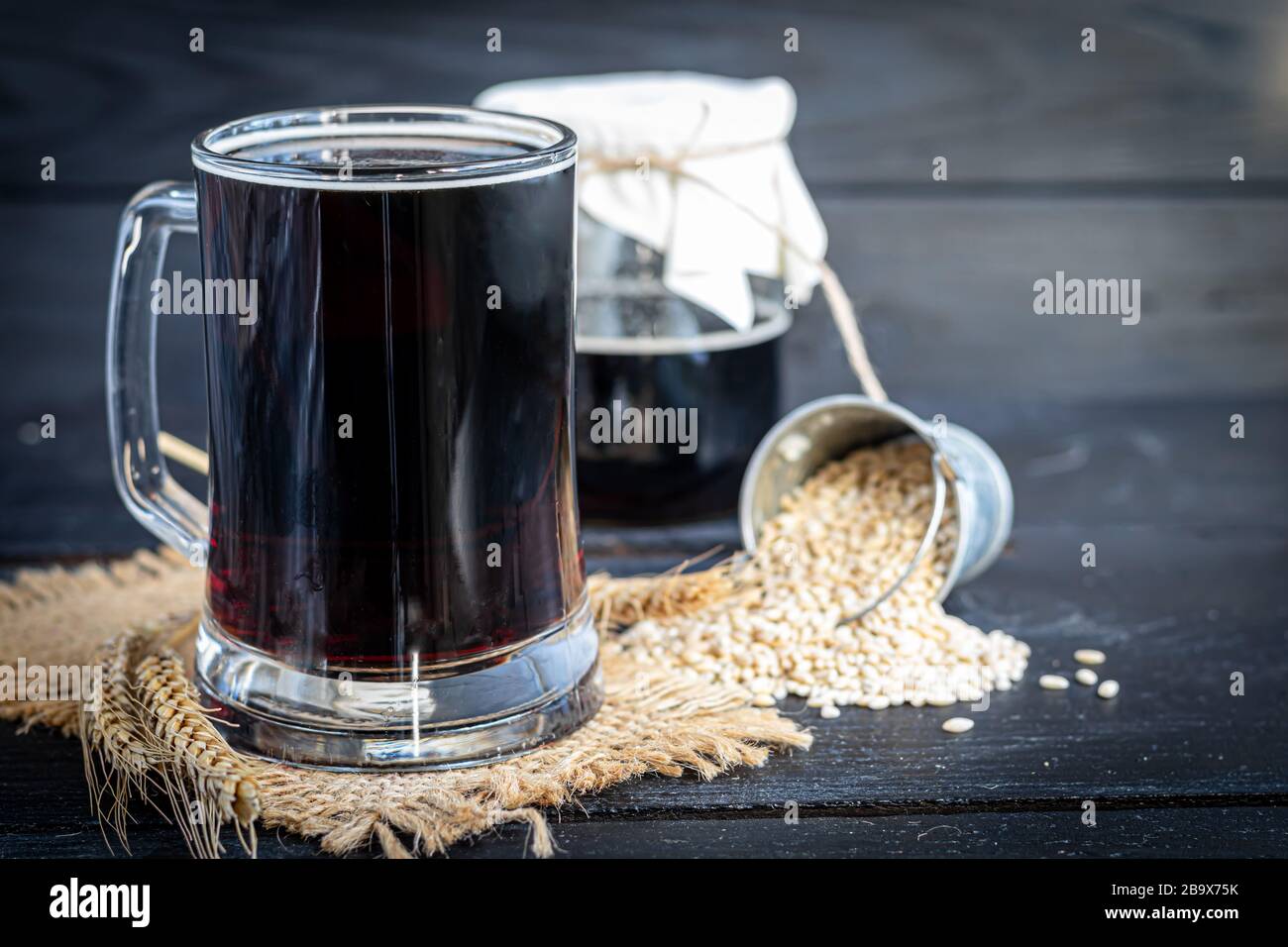 Hausgemachtes dunkles Bier in Flaschen und ein Glas auf dunklem Hintergrund. In der Nähe liegen Gerstenkörner und Weizenkörner. Dunkler Hintergrund. Kopierbereich Stockfoto