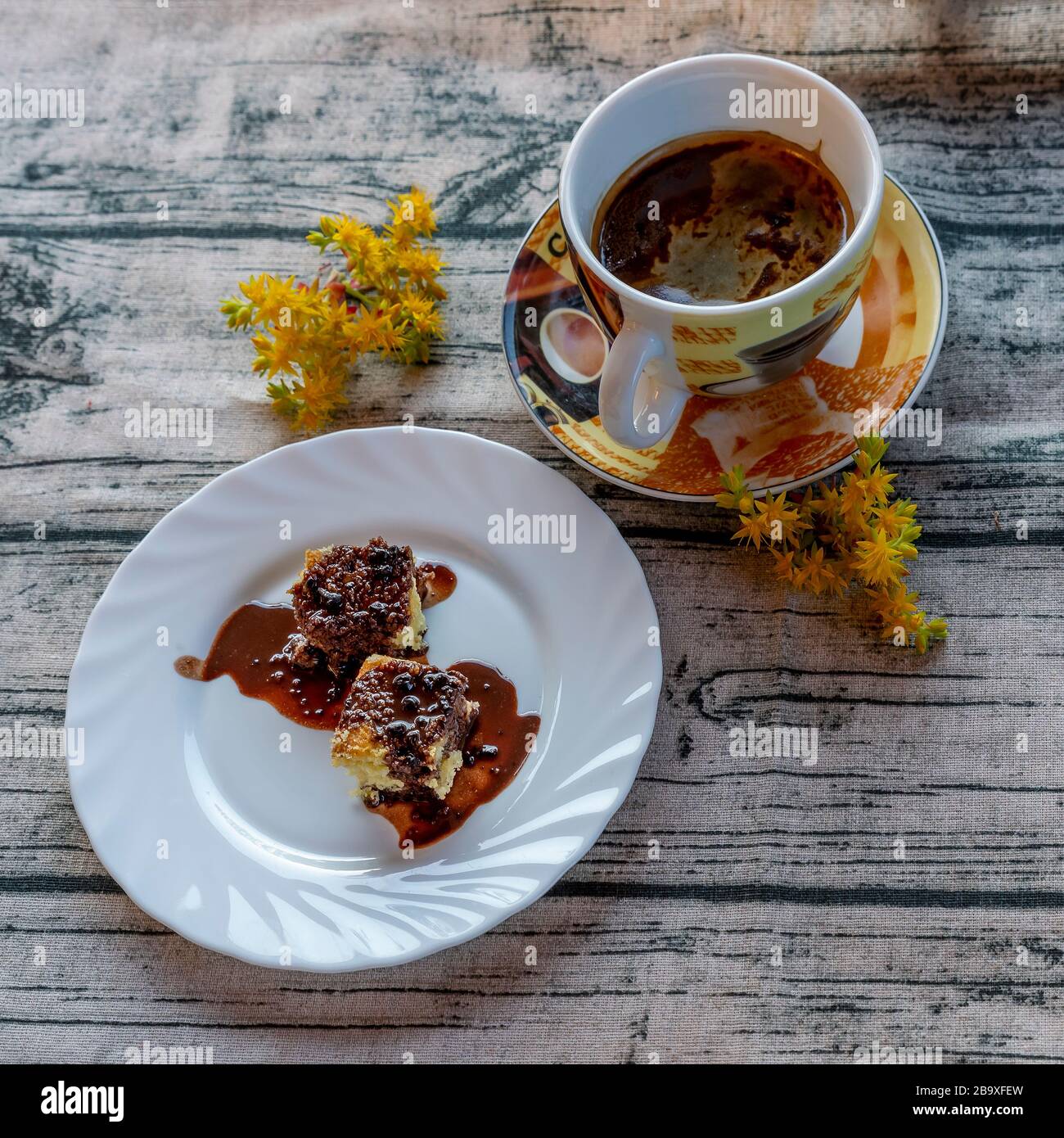 Das köstliche traditionelle italienische Dessert namens Mimosa, das mit flüssiger Schokolade mit einer Tasse Gerstenkaffee und gelben Sedumblumen bedeckt ist Stockfoto