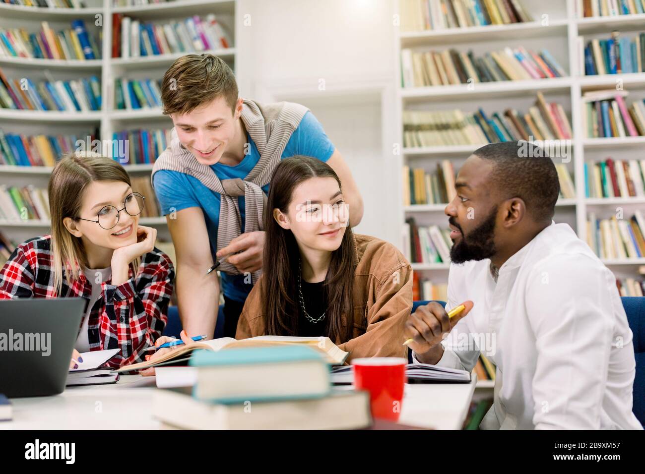 Lächelnde, fröhliche moderne multirassische Studenten, die während ihrer Vorbereitung auf Prüfungen oder praktische Klassen in der Bibliothek sitzen und Bücher lesen, teilen Stockfoto