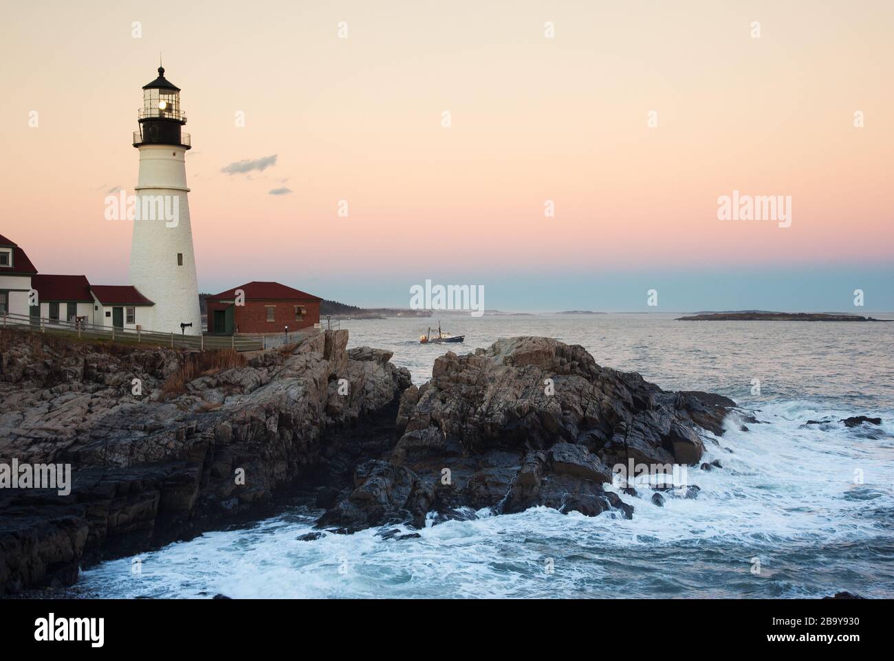 Der Leuchtturm von Portland Head führt das Fischereischiff aus der Casco Bay, während die Sonne in Maine untergeht. Der Turm scheint hell über die felsige Küste. Stockfoto