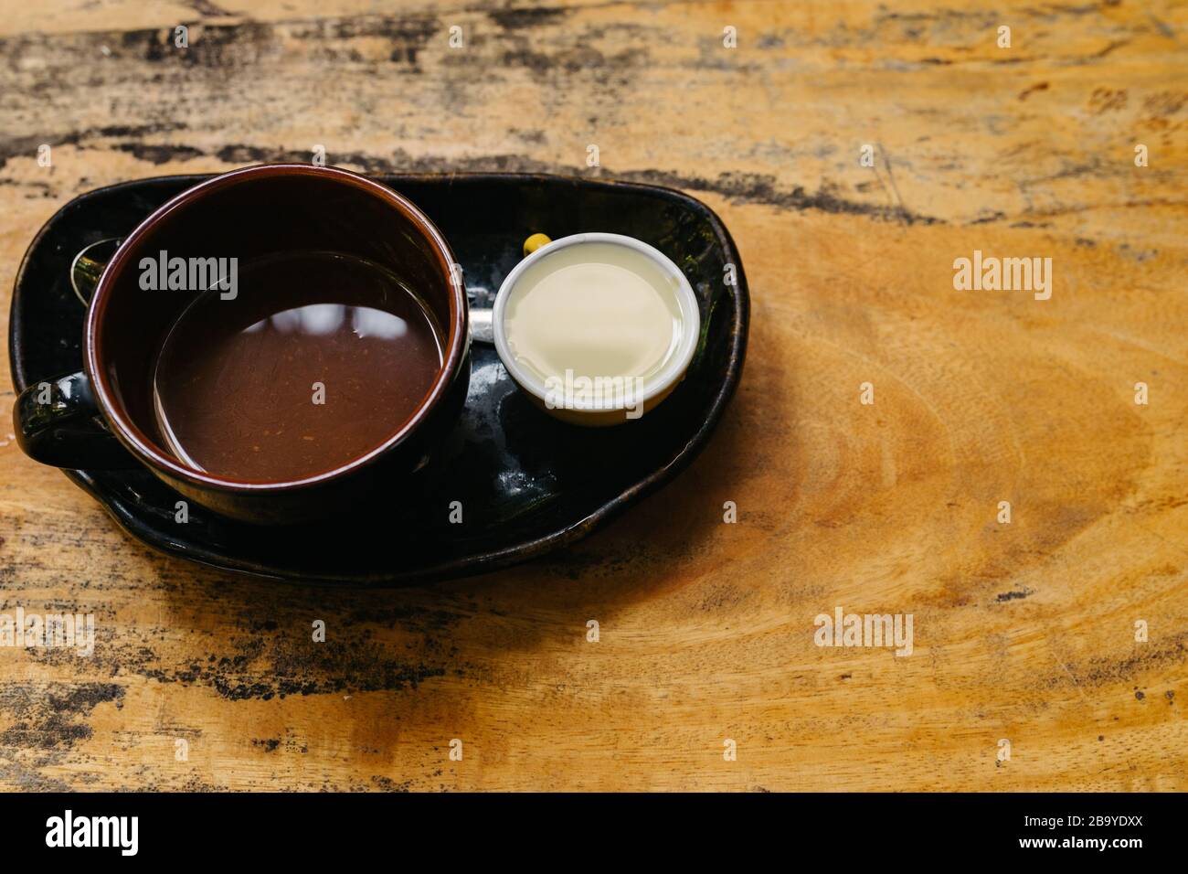 Eine Tasse heißen Espresso in einer dunklen Tasse auf einer schwarzen Untertasse mit Milch separat. Aromatischer Kaffee steht auf einem alten braunen Holztisch. Ort kopieren. Ein guter ST Stockfoto