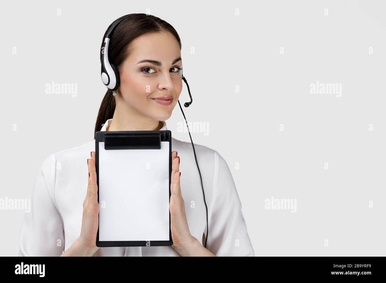 Schöne lächelnde Frau Beraterin des Callcenters in Kopfhörern, die die Zwischenablage mit einem leeren weißen Blatt auf grauem Hintergrund halten. Weibliche Kundenbetreuerin mit Headset Stockfoto