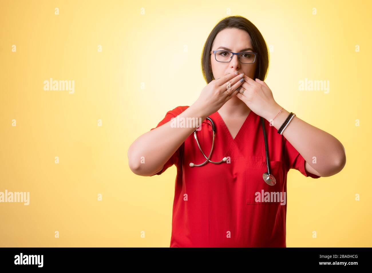 Porträt einer schönen Frau Doktor mit Stethoskop mit roten Schruppen, bedeckt Mund wie Mute-Konzept, posiert auf einem gelb isolierten Hintergrund. Stockfoto