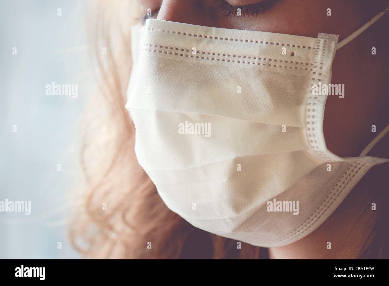Detail der kaukasischen Frau, die eine weiße medizinische Gesichtsmaske trägt. Frau weint, geschlossene Augen. Fokussieren Sie den vorderen Teil des Gesichts und den verschwommenen Hintergrund. Coronavirus, COVID-19 Quarantäne. Pflegekonzept. Stockfoto
