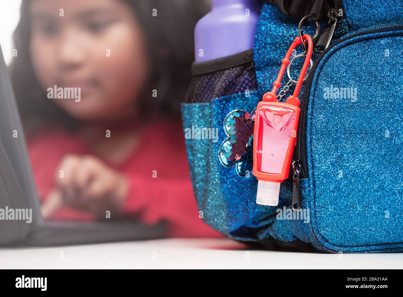 Gel-Hanitisierer, der an einem Rucksack mit Metall-Wasserflasche auf einem Schreibtisch befestigt ist, und der Student verwendet im Hintergrund einen Laptop. Stockfoto