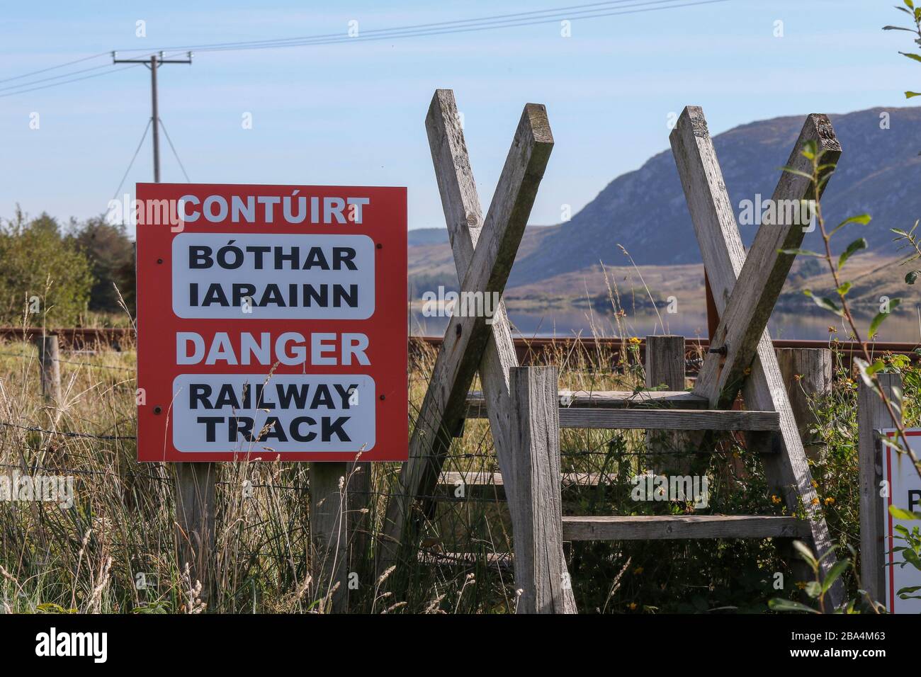 Ein rot-weißes Warnschild in irischer und englischer Sprache neben einem Holzstile an der Fintown Railway Track in einer Gaeltacht Gegend der Grafschaft Donegal, Irland. Stockfoto