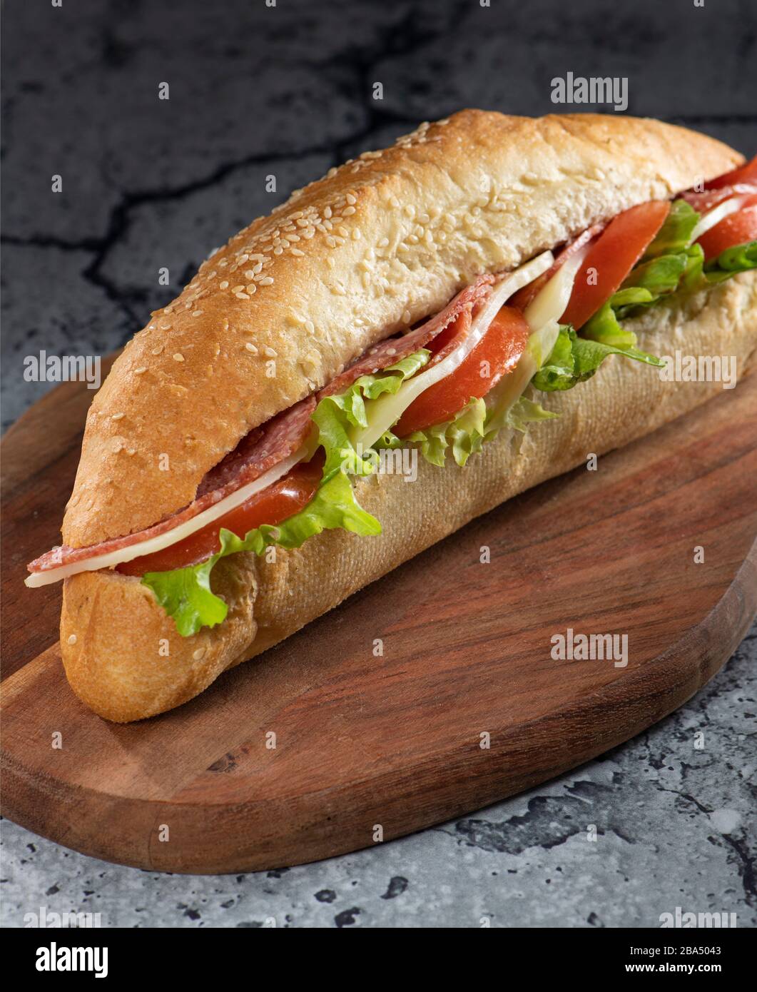 Ein Baguette-Sandwich auf einem Schneidebrett mit Tomaten, Cheddar, Salami, Salat, Mayonnaise Stockfoto