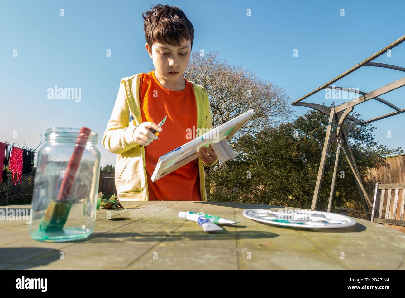 Ein kleiner Junge, der an einem sonnigen Tag ein Gemälde auf einem Tisch im Hintergarten macht. Stockfoto