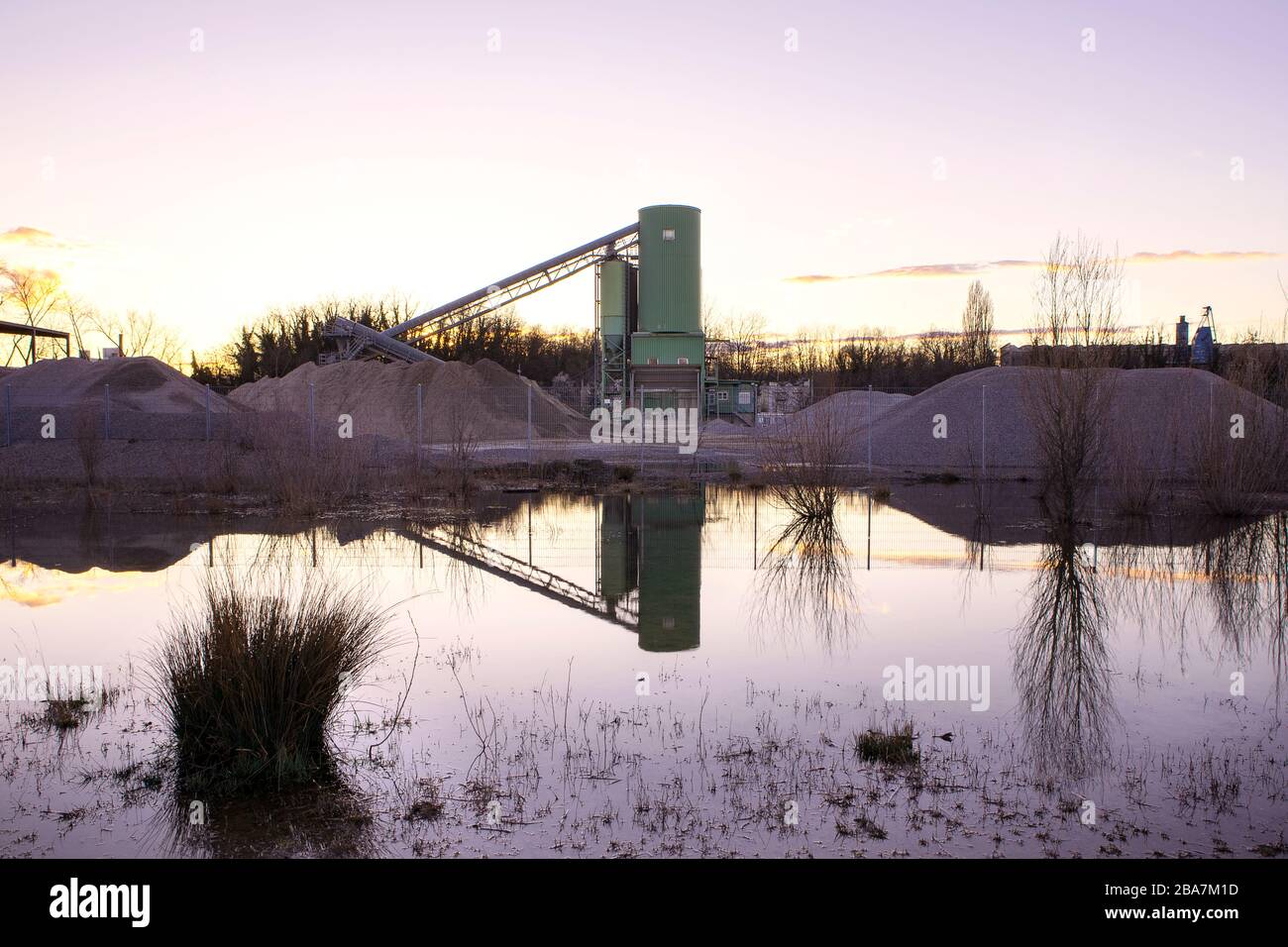 Förderband über Schottergrube bei Sonnenuntergang. Industrielle Landschaft, Gebäude reflektieren im Wasser. Feuchtgebiet Rheinfluss, Deutschland. Stockfoto