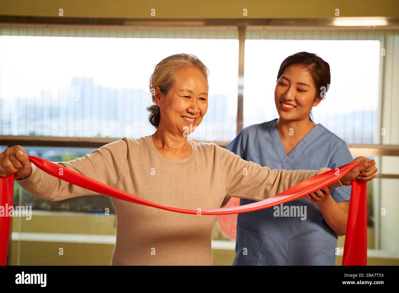 Glückliche ältere asiatische Frau, die mit einer Widerstandsband trainiert, die von einem physischen Therapeuten im Reha-Zentrum geleitet wird Stockfoto