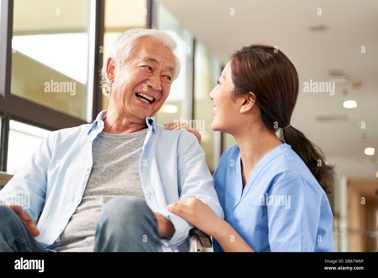 Junge freundliche asiatische Pflegekräfte unterhalten sich im Flur des Pflegeheims mit einem glücklichen Senioren-Mann Stockfoto