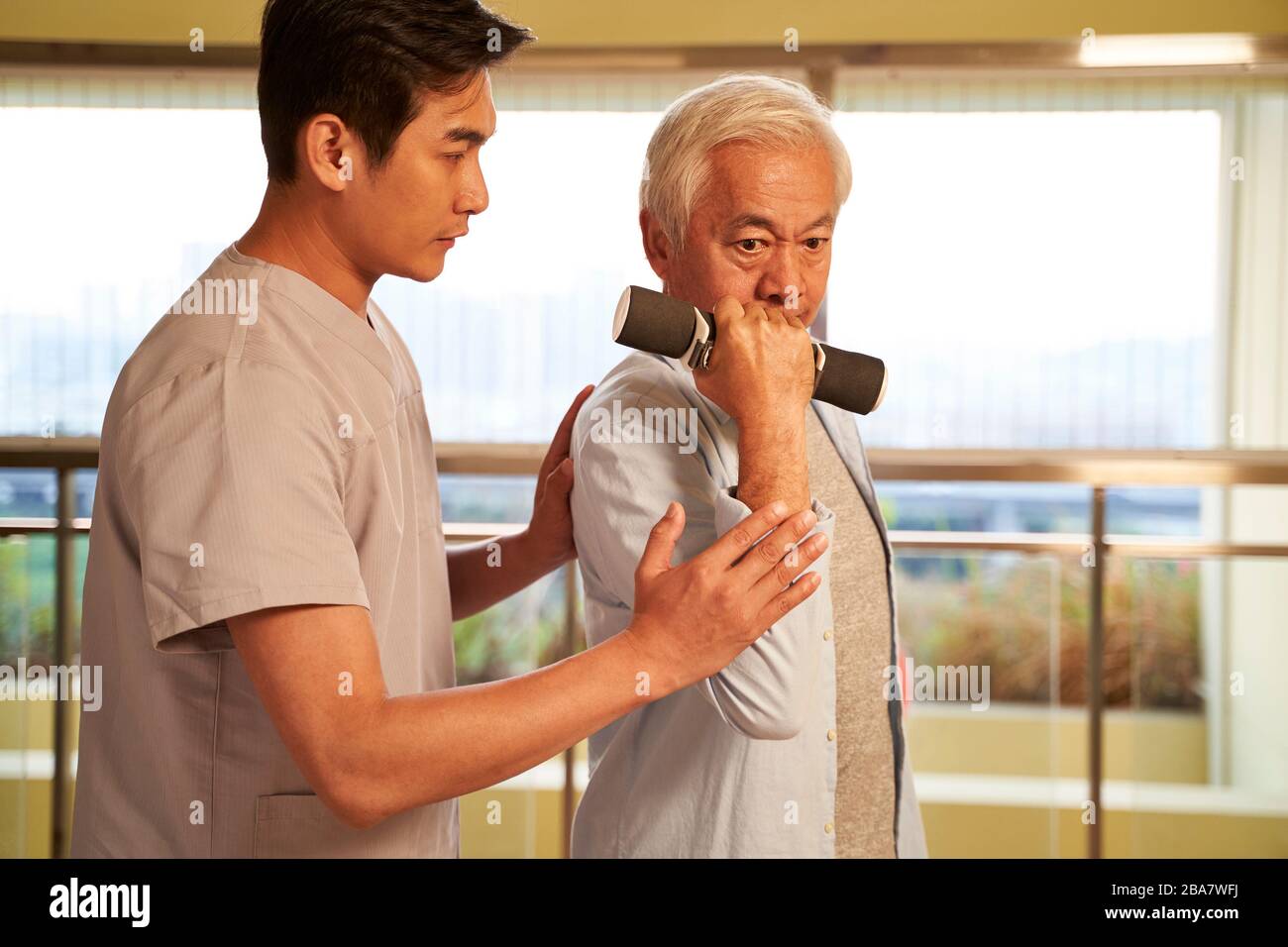 Leitender asiatischer Mann, der mit Hanteln trainiert, die von einem physischen Therapeuten im Rehabilitationszentrum geleitet werden Stockfoto