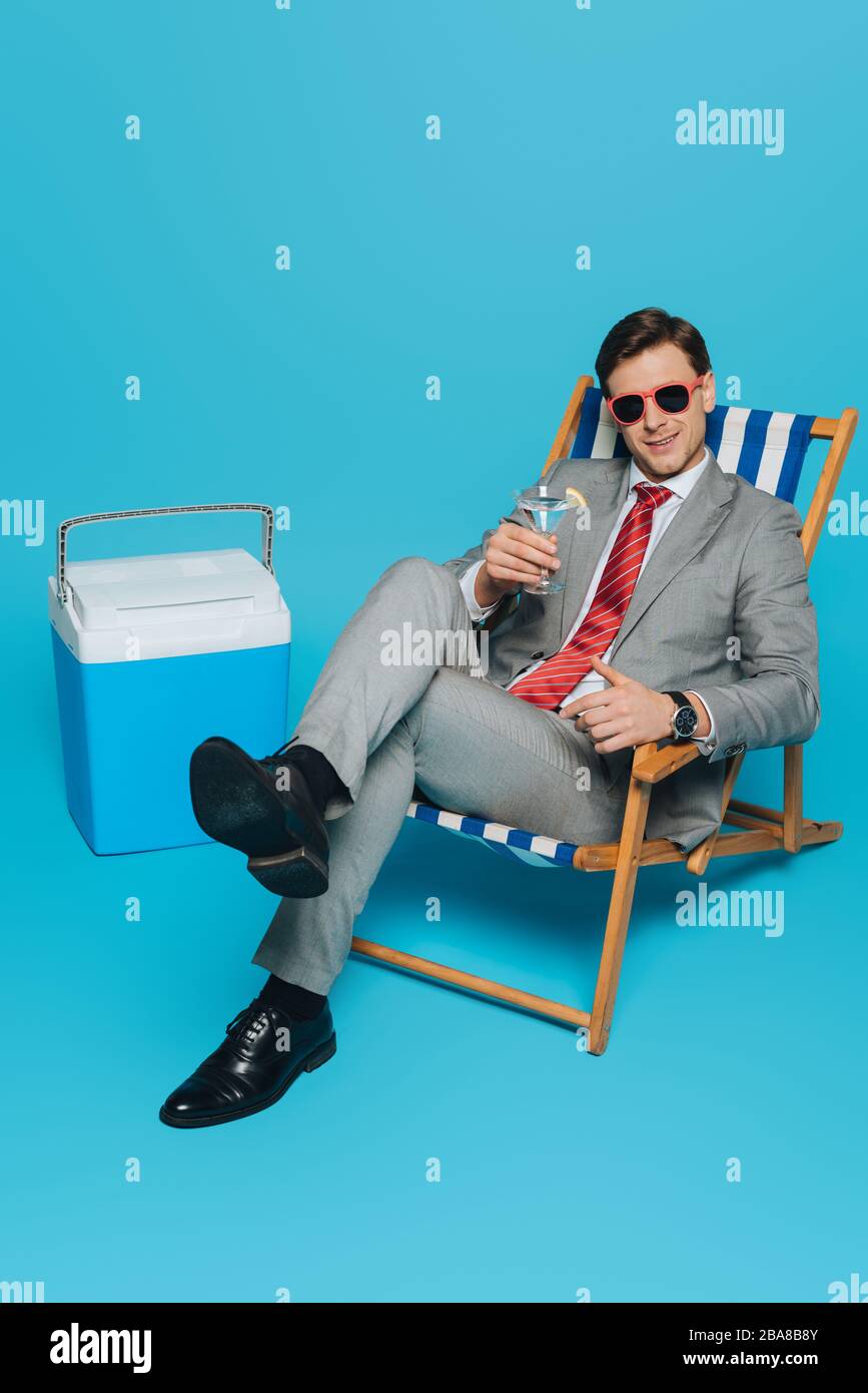 Lächelnder Geschäftsmann in einer Sonnenbrille auf dem Liegestuhl mit einem Glas Cocktail in der Nähe eines tragbaren Kühlschranks auf blauem Hintergrund Stockfoto