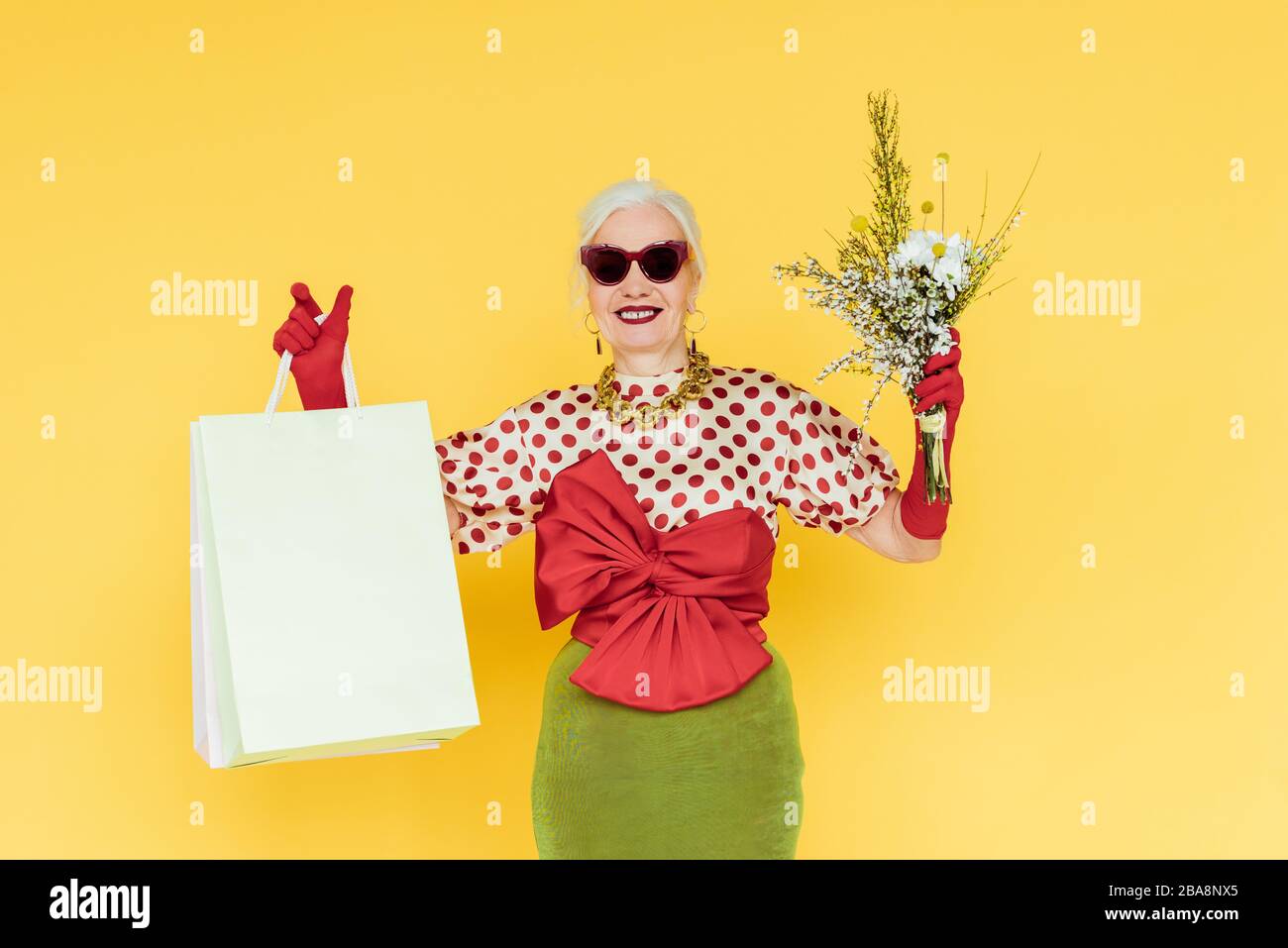 Stilvoll lächelnde Seniorin, die Wildblumen und Einkaufstaschen auf gelbem Hintergrund hält Stockfoto