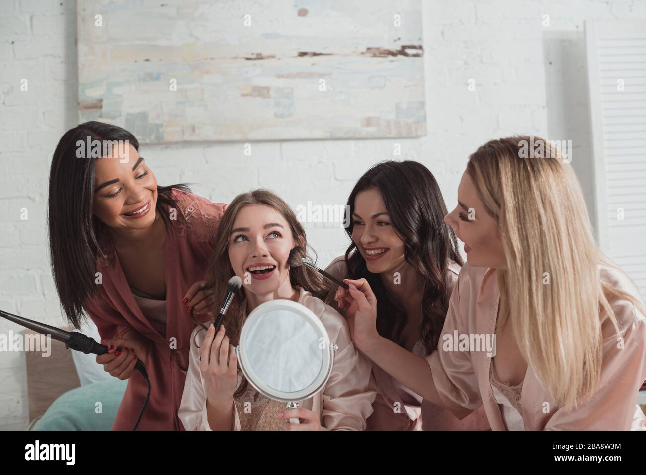 Multikulturelle Frauen, die auf der Junggesellen-Party mit Kosmetikbürsten und Spiegel und Eisenstab aufmachen Stockfoto