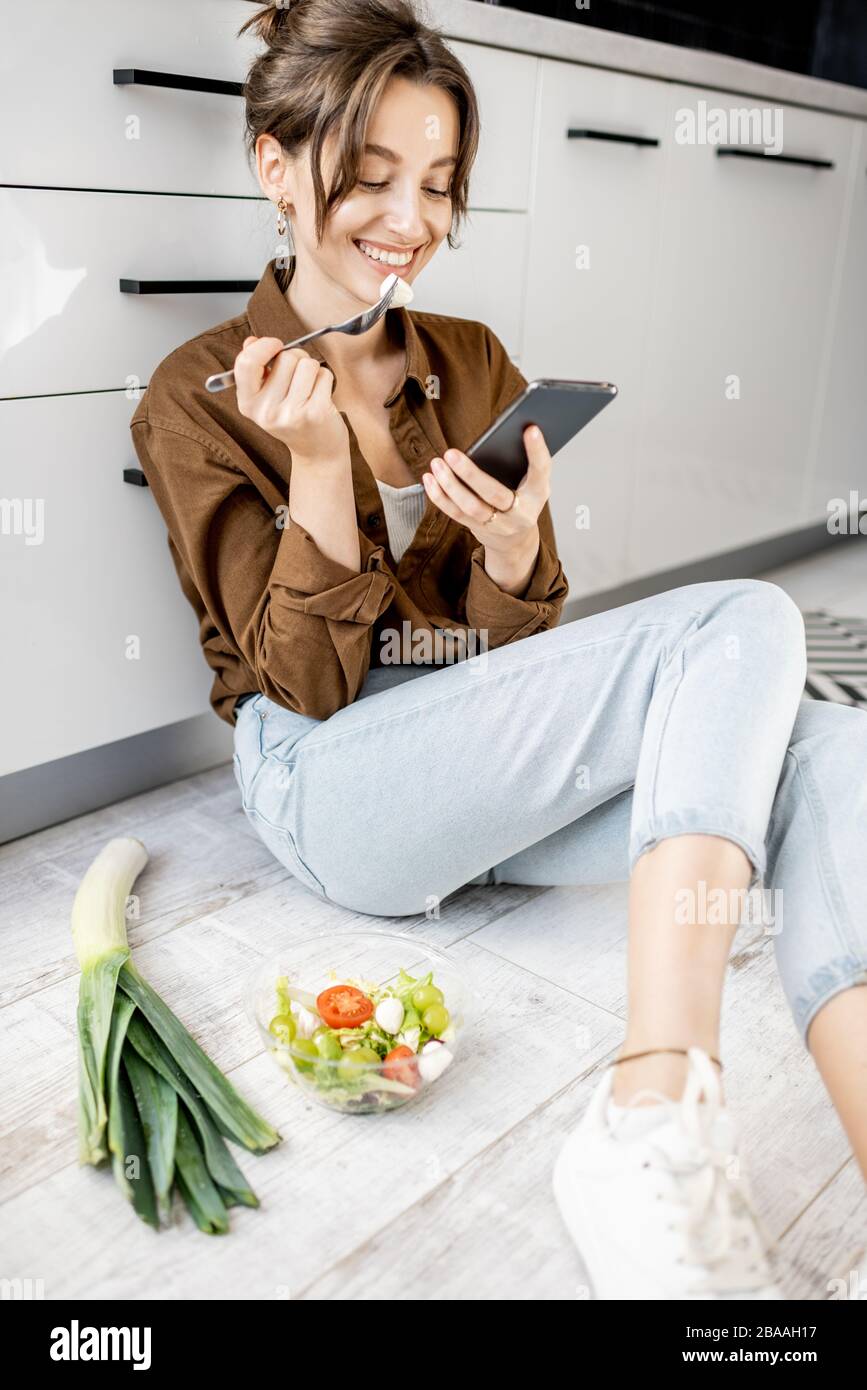 Junge Frau sitzt mit Telefon auf dem Küchenboden und isst Salat, während sie zu Hause entspannt ist. Gesundes Essverhalten und Lifestyle-Konzept Stockfoto