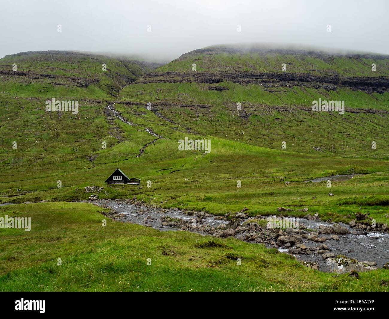 Färöer. Grüne Felder aus Gras, von Flüssen und kleinen Wasserfällen geschnitten. Raues Wetter. Wolken über Hügeln. Stockfoto