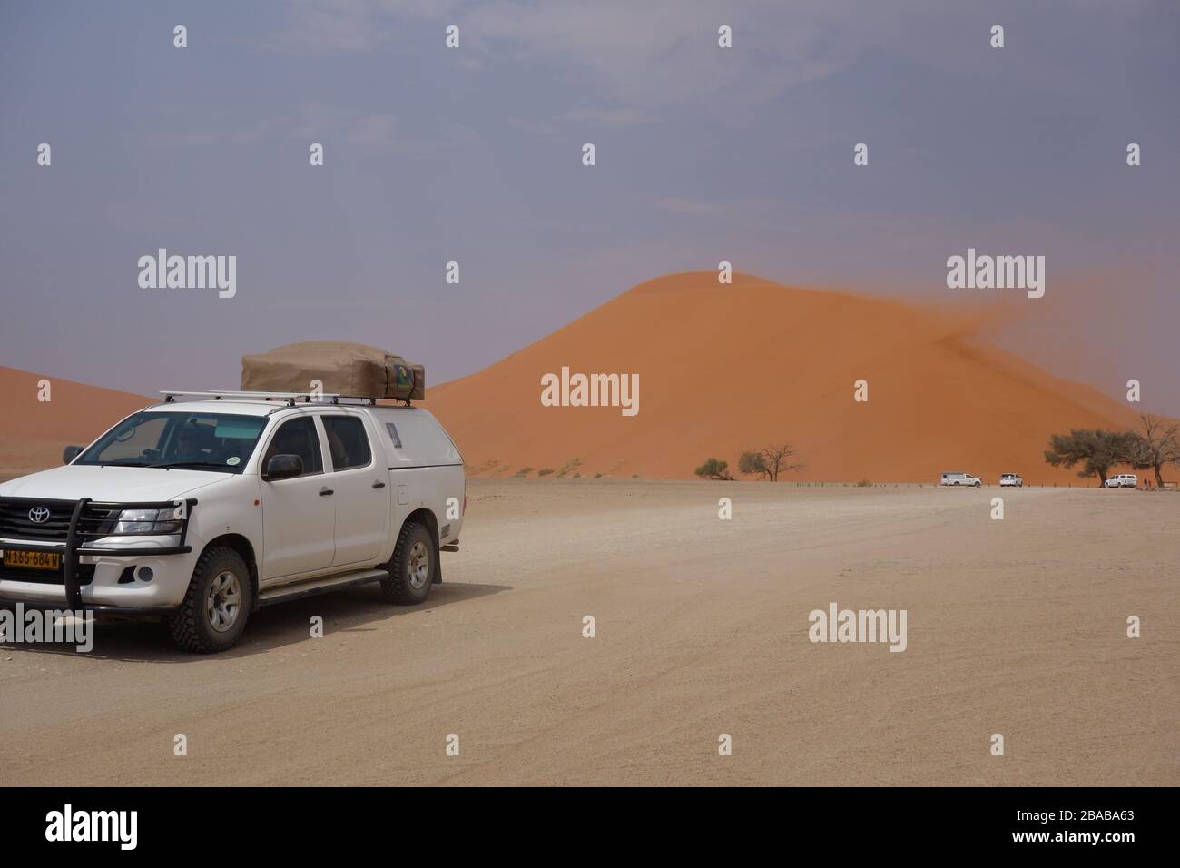 Weißer Geländewagen mit Geländewagen und Geländewagen, mit Touristengruppe, die vor der roten Sanddüne/Sanddüne in der Wüste Sossusvlei in Namibia mit Sandsturmwind fährt Stockfoto