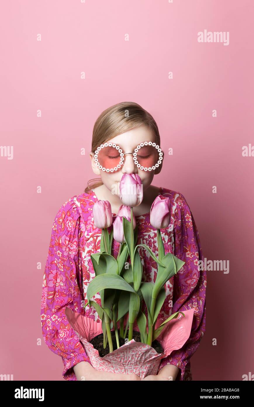Blonde Tween Mädchen in Sonnenbrille riecht rosa Tulpen auf rosa Hintergrund Stockfoto
