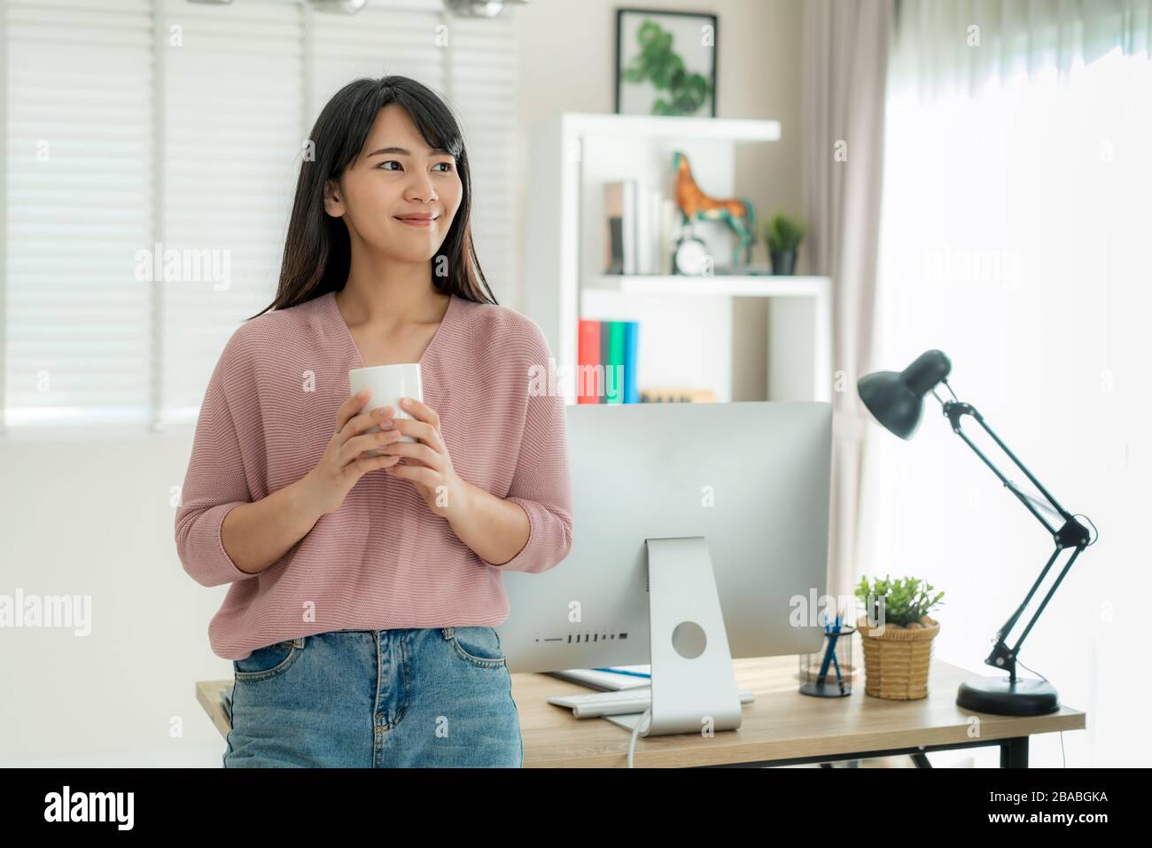 Asiatische schöne junge Frau arbeitet von zu Hause aus an Computer und trinkt Kaffee, während sie sich von ihrer Arbeit im Wohnzimmer zu Hause entspannen kann. Sozial di Stockfoto