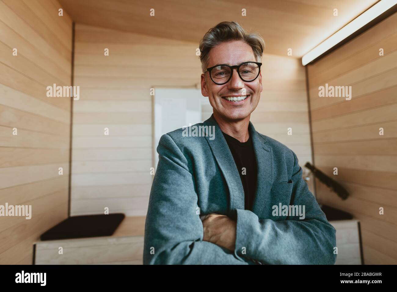 Mitteladulter Geschäftsmann, der mit seinen im Amt gekreuzten Armen steht. Reifer Manager in der Formalkleidung mit Blick auf die Kamera und lächelnd. Stockfoto