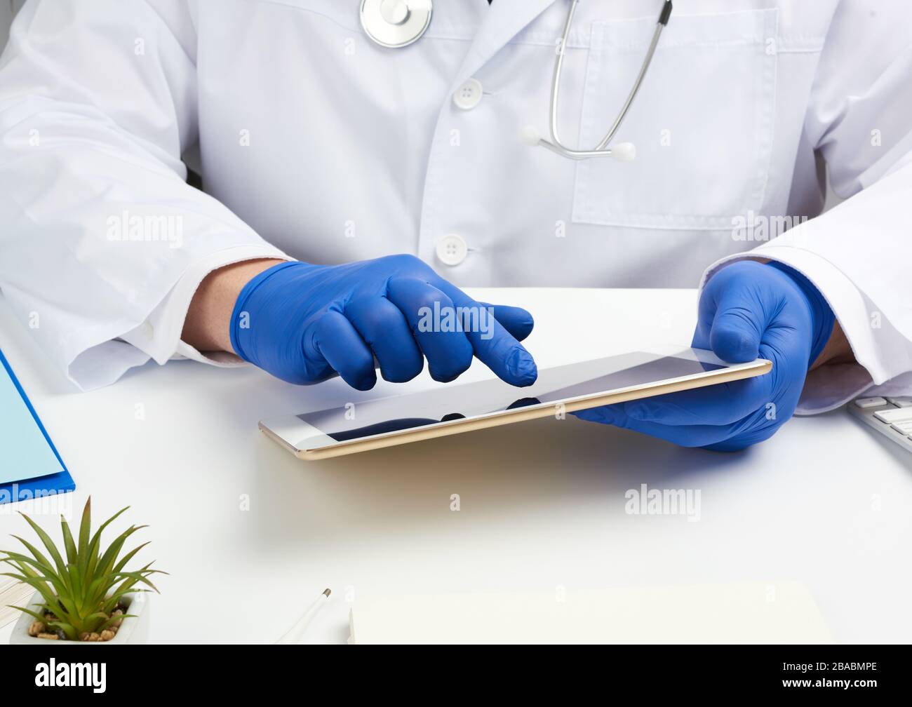 Doktor in einem weißen medizinischen Mantel sitzt an einem Tisch und hält eine elektronische Tablette in der Hand, berührt den Bildschirm mit der rechten Hand, Arztpraxis, di Stockfoto