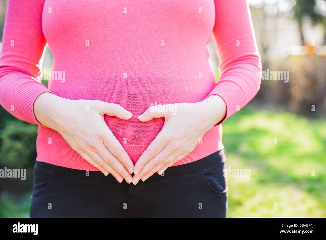 Nahaufnahme einer schwangeren Frau, die an einem sonnigen Frühlingnachmittag draußen steht und ihre Hände in Form eines Herzens am Bauch hält Stockfoto