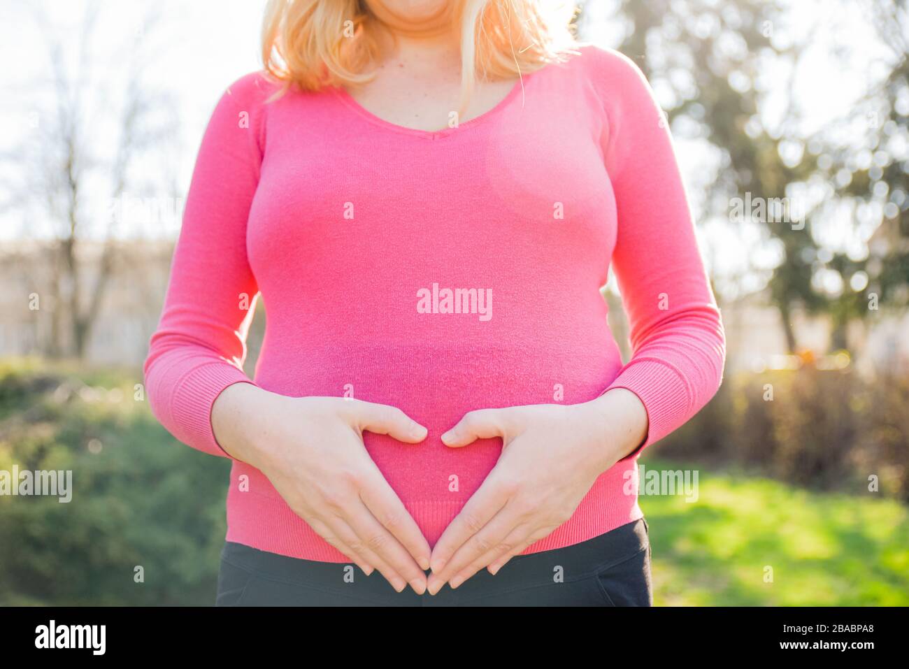 Schwangere Dame in rosafarbenem Hemd, die an einem sonnigen Frühlingnachmittag draußen steht und ihre Hände am Bauch hält Stockfoto