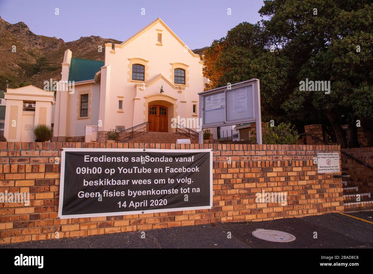 KAPSTADT, SÜDAFRIKA - 26. MÄRZ 2020: Die NG Kerk (Kirche) in der Gordons Bay mit einem Schild an der Wand, das besagt, dass alle Gottesdienste ausgesetzt sind und die Anbeter auf YouTube und Facebook folgen können, während die Sonne am Abend vor einem 21-tägigen nationalen Lockdown der südafrikanischen Regierung zur Bekämpfung der untergeht Ausbreitung des Covid-19 oder Coronavirus. Foto von Roger Sedres Stockfoto