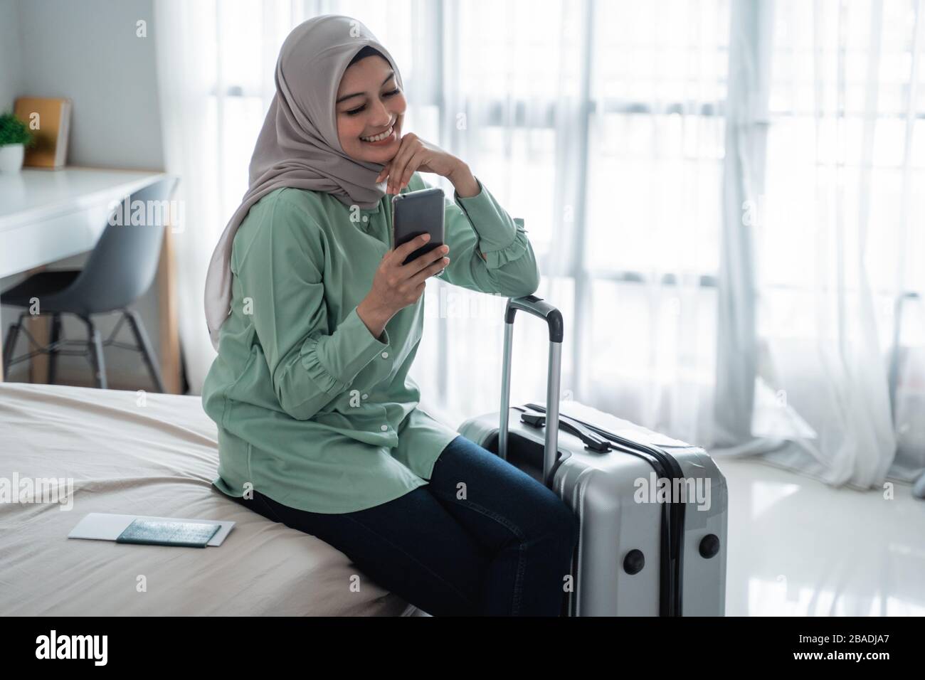Die verschleierte Frau saß auf dem Bett und hielt ihr Smartphone und ihren Koffer, bevor sie in den Urlaub ging Stockfoto