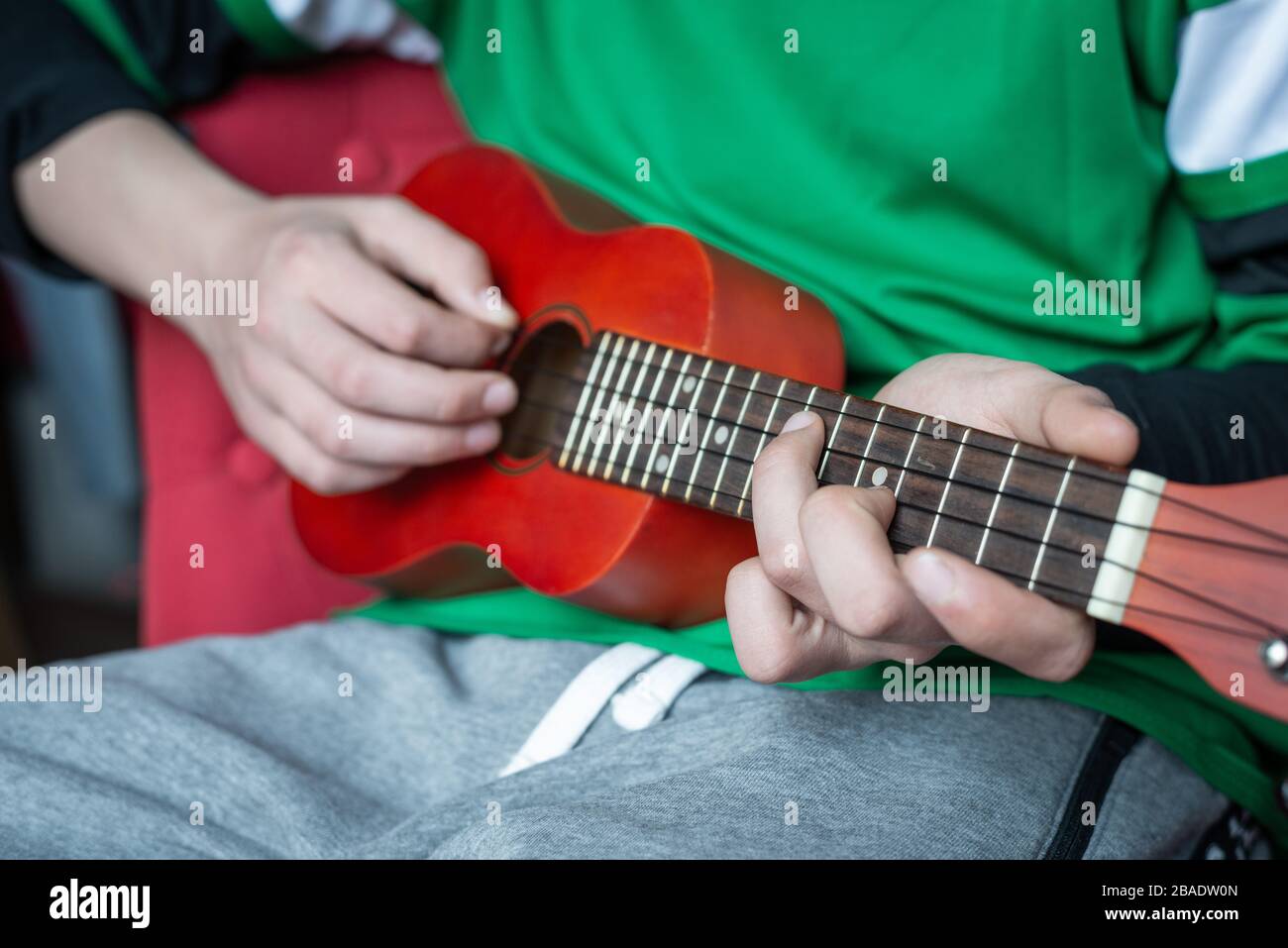 Kleiner Junge, der zu Hause eine rote Ukelele spielt, Nahaufnahme der Hände. Stockfoto