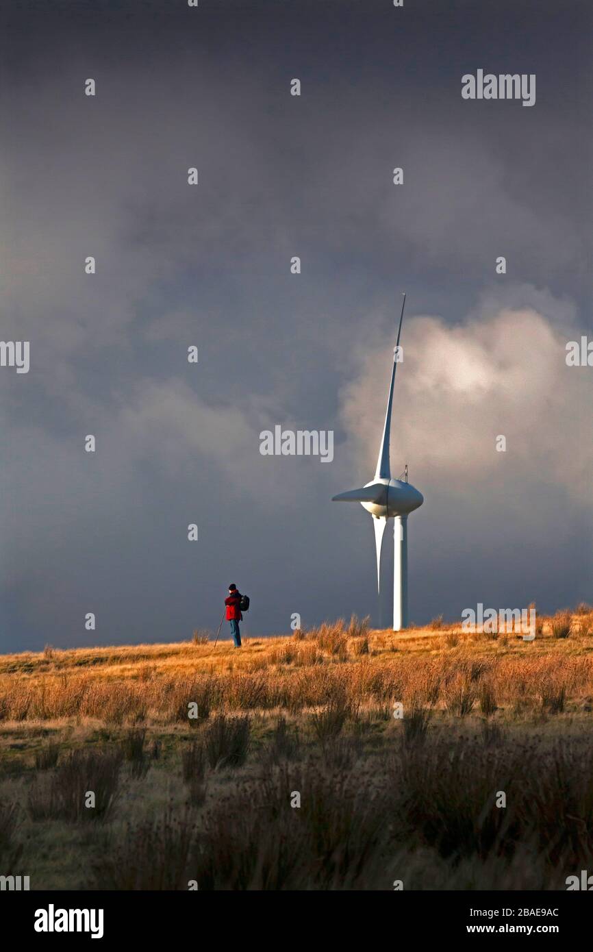 Einsamer Bergwanderer auf dem Hügelkamm mit Windturbine und stürmischem Himmel mit dramatischem Licht, Wales, Großbritannien Stockfoto