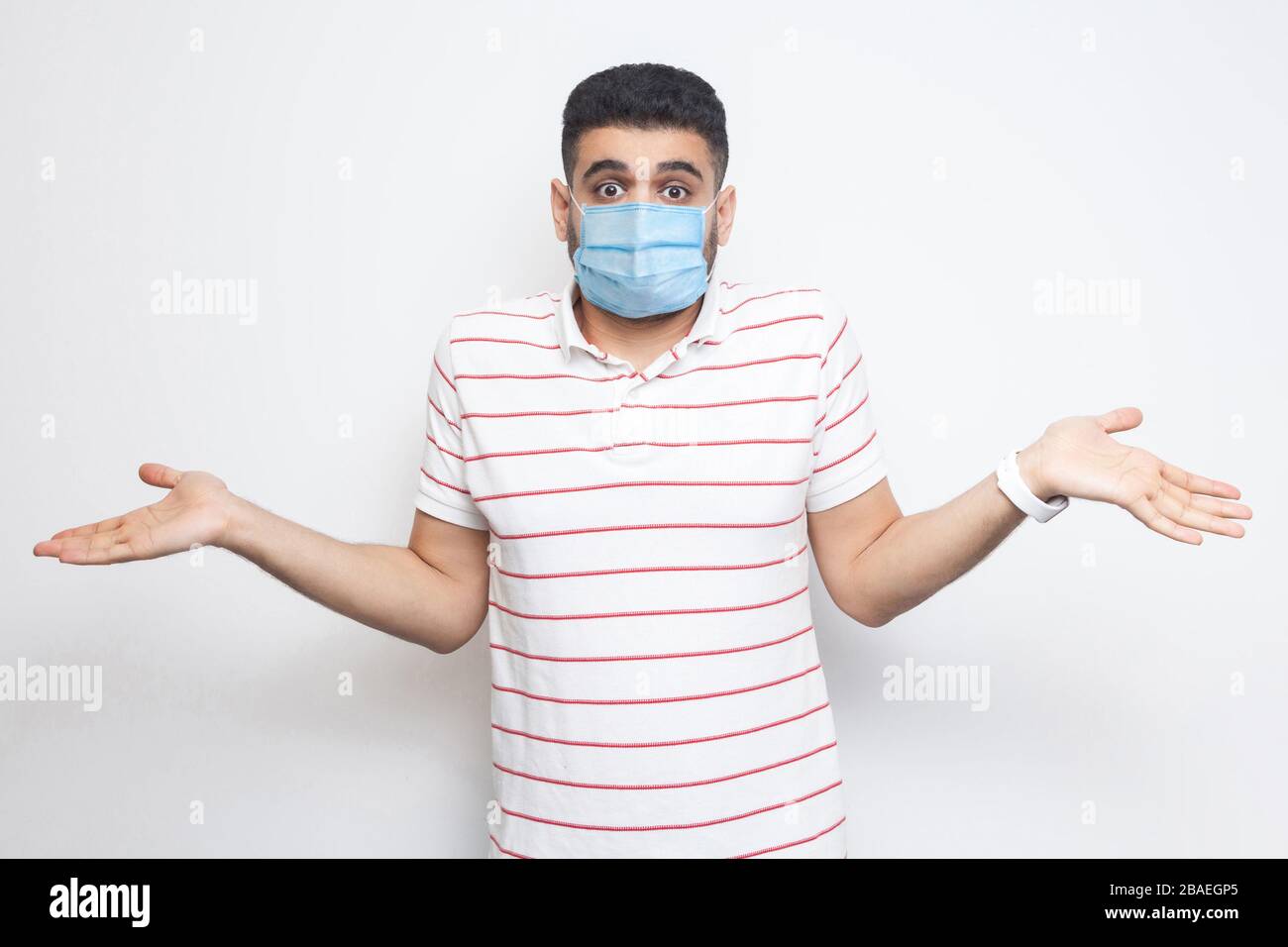 Ich weiß es nicht. Portrait des verwirrten jungen Mannes mit chirurgischer medizinischer Maske im gestreiften T-Shirt, das mit erhobenen Armen steht und nicht weiß, was zu tun ist. Innen Stockfoto