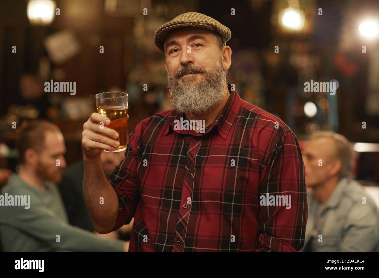 Reifer bärtiger Mann in legerer Kleidung, der ein Glas Bier hält und mit seinen Freunden die Kamera betrachtet, während er in der Kneipe ruht Stockfoto