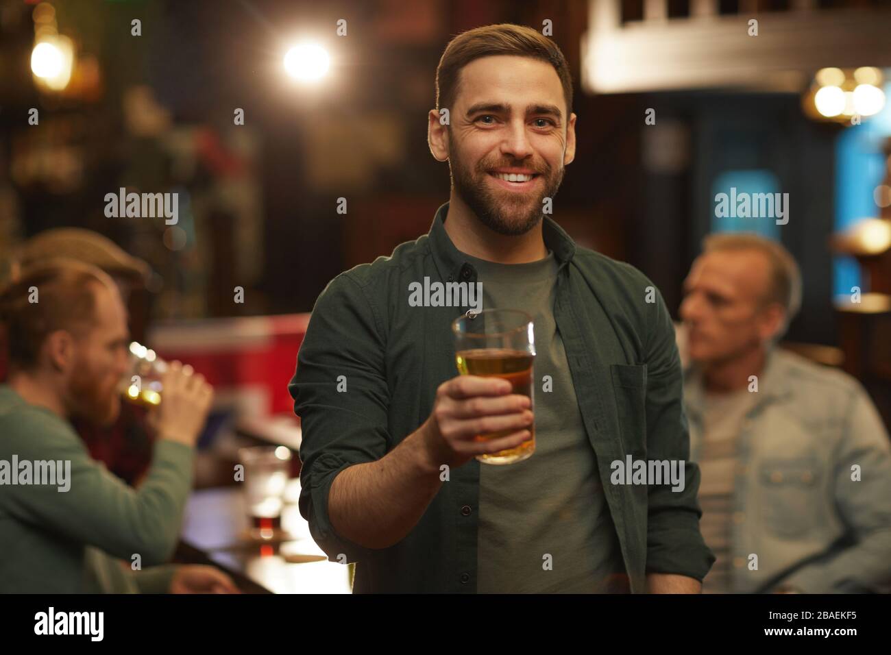 Porträt des jungen Mannes in legerer Kleidung lächelt an der Kamera und trinkt mit seinen Freunden in der Kneipe Bier Stockfoto
