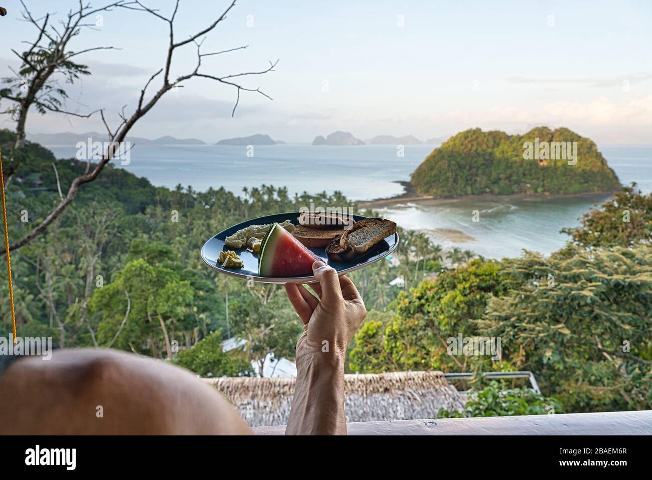 Mann mit gesundem kontinentalen Frühstück vor einem paradiesischen Strand mit Blick auf El Nido, Philippinen. Wassermelone, Rührei, Toast und Butter Stockfoto