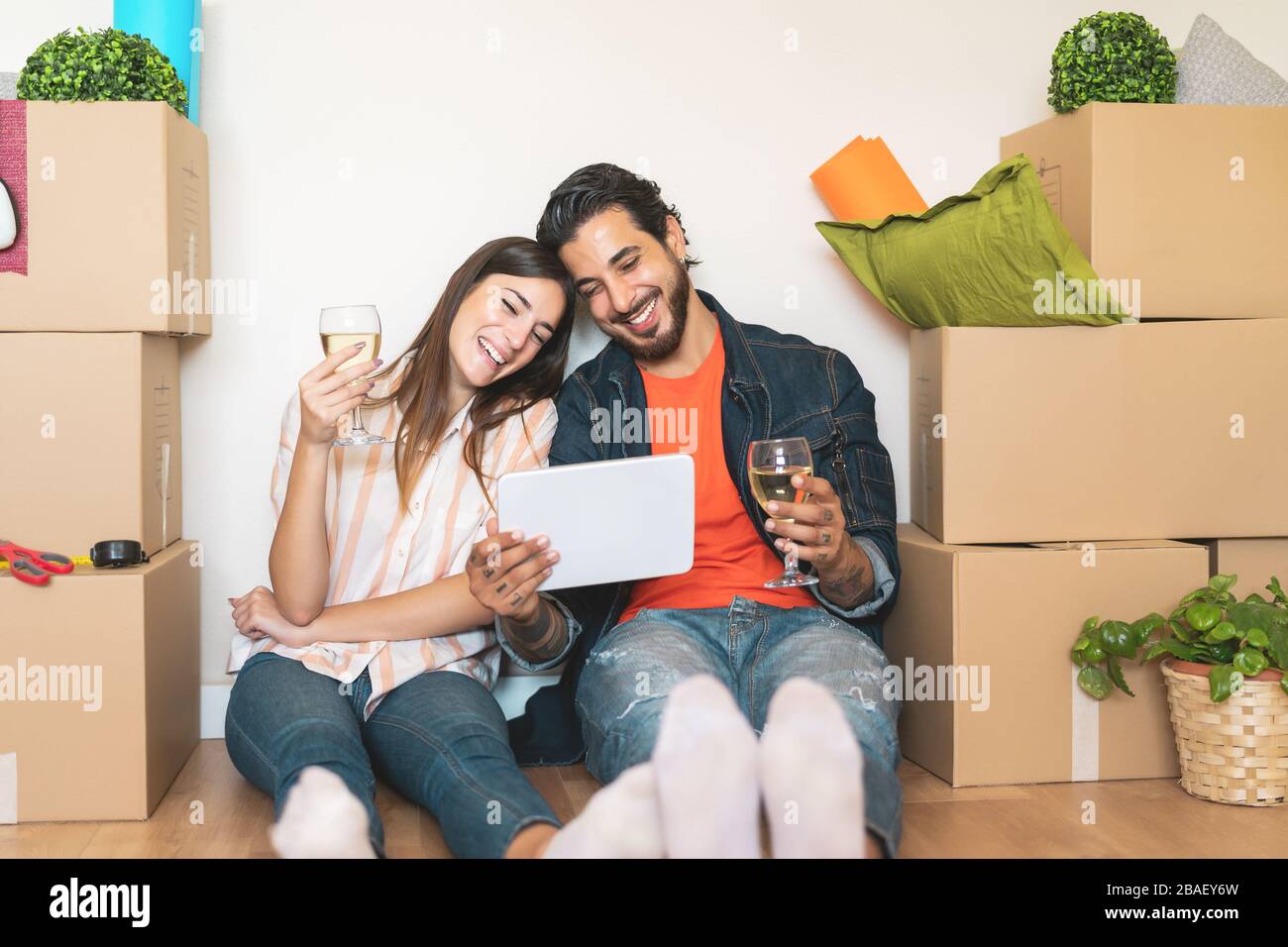 Glückliches junges Paar zieht in ein neues Haus - junge Liebhaber haben Spaß mit Tablet-PC-Shopping online sitzend neben Karton Stockfoto