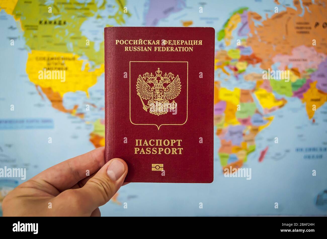 Hand, die den Reisepass Russlands gegen den bunten Weltkarten-Atlas hält. Russisches Passkonzept, Reisekonzept russischer Touristen. Stockfoto
